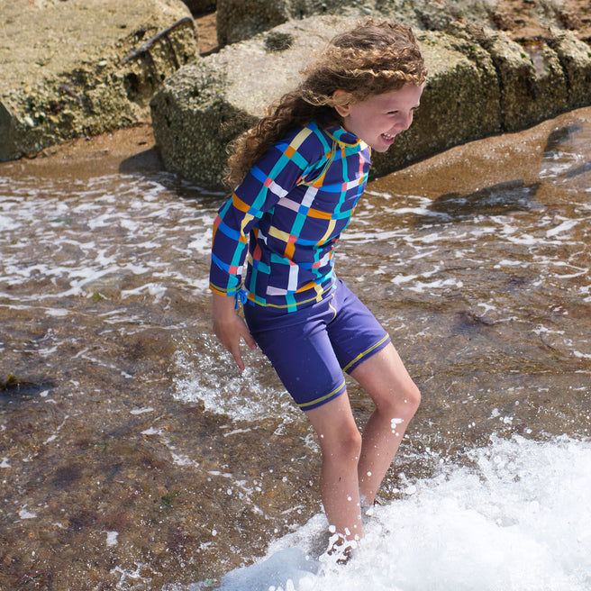 A girl plays in the surf wearing a UV Protective Surf Set Multi Check, smiling as the waves crash around her legs.
